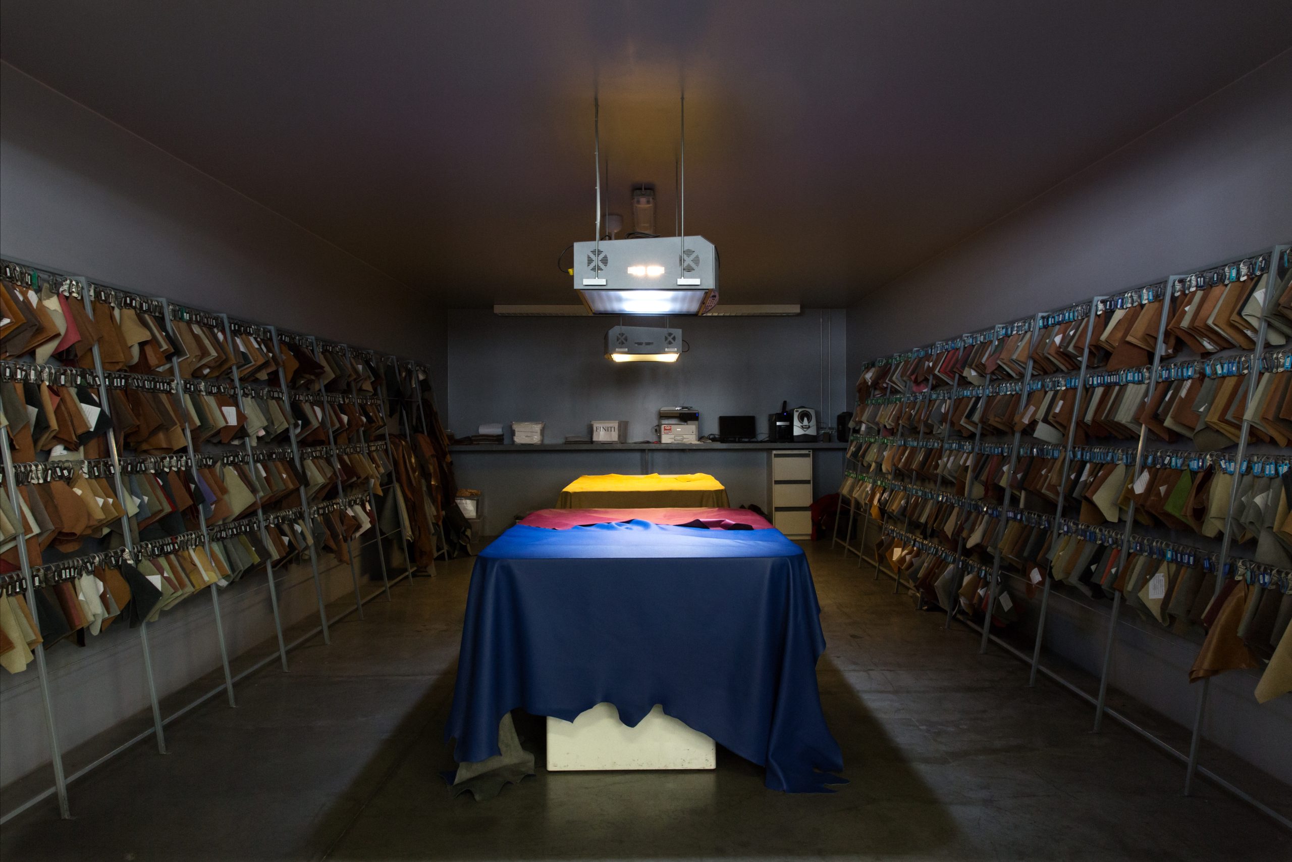 Leather sample room with rows of leather swatches displayed on racks along both walls and a central table under overhead lighting.
